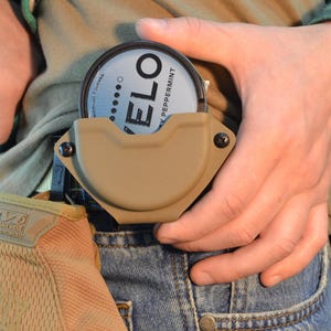 May include: Tan-colored holster holding a round container with the text "VELO" and "DARK PEPPERMINT". The holster is attached to a belt, and the person is wearing a green and black plaid shirt and blue jeans.