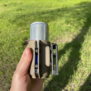May include: A tan-colored can holder with a black clip is shown. The holder is designed to secure a silver can, with a blue object visible inside. The background is a blurred green grassy area.
