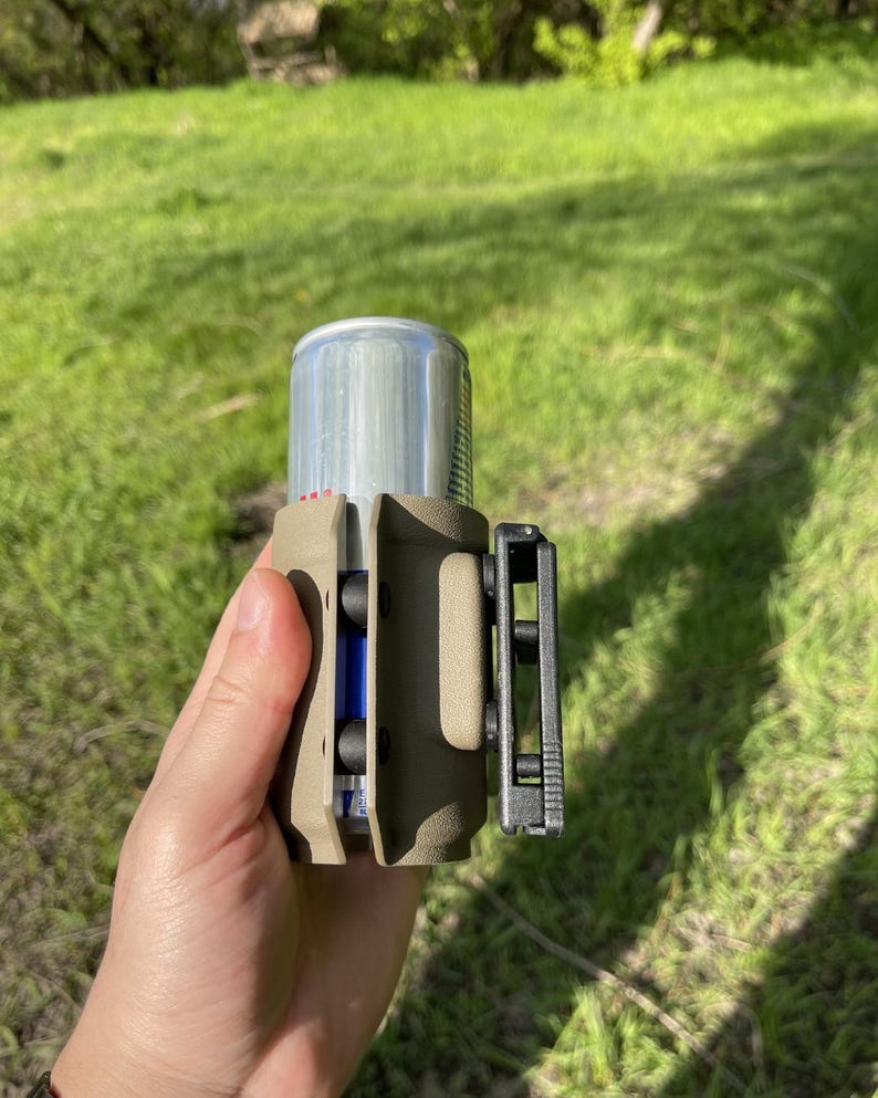 May include: A tan-colored can holder with a black clip is shown. The holder is designed to secure a silver can, with a blue object visible inside. The background is a blurred green grassy area.