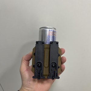 May include: A tan and black tactical can holder with a silver Red Bull energy drink can. The holder has two black clips and is designed to securely hold a standard-sized beverage can. The holder is held in a hand.