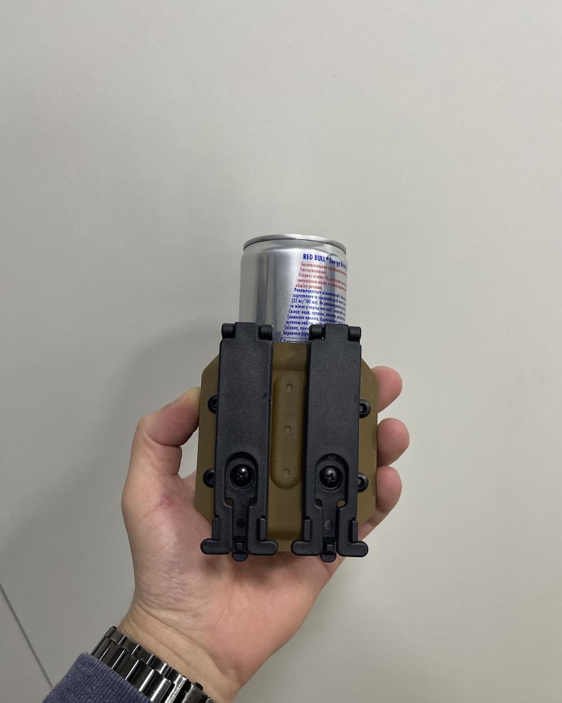 May include: A tan and black tactical can holder with a silver Red Bull energy drink can. The holder has two black clips and is designed to securely hold a standard-sized beverage can. The holder is held in a hand.