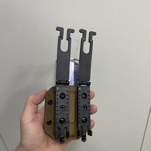 May include: A black and tan tactical holster with two black belt clips. The holster has multiple mounting holes and is designed to hold a cylindrical object, possibly a magazine or a similar item.