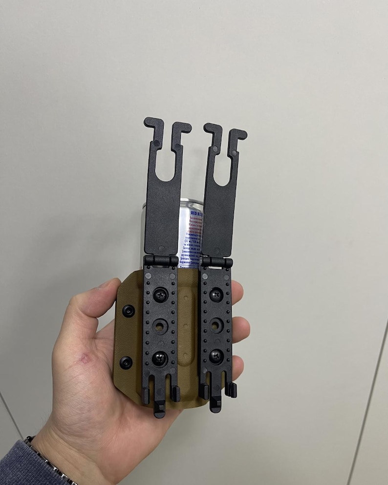 May include: A black and tan tactical holster with two black belt clips. The holster has multiple mounting holes and is designed to hold a cylindrical object, possibly a magazine or a similar item.