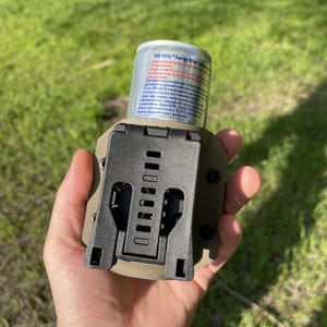 May include: A tactical beverage holder in olive green and black, designed to securely hold a Red Bull energy drink can. The holder features a sturdy, adjustable clip and is held in a hand against a blurred green background.