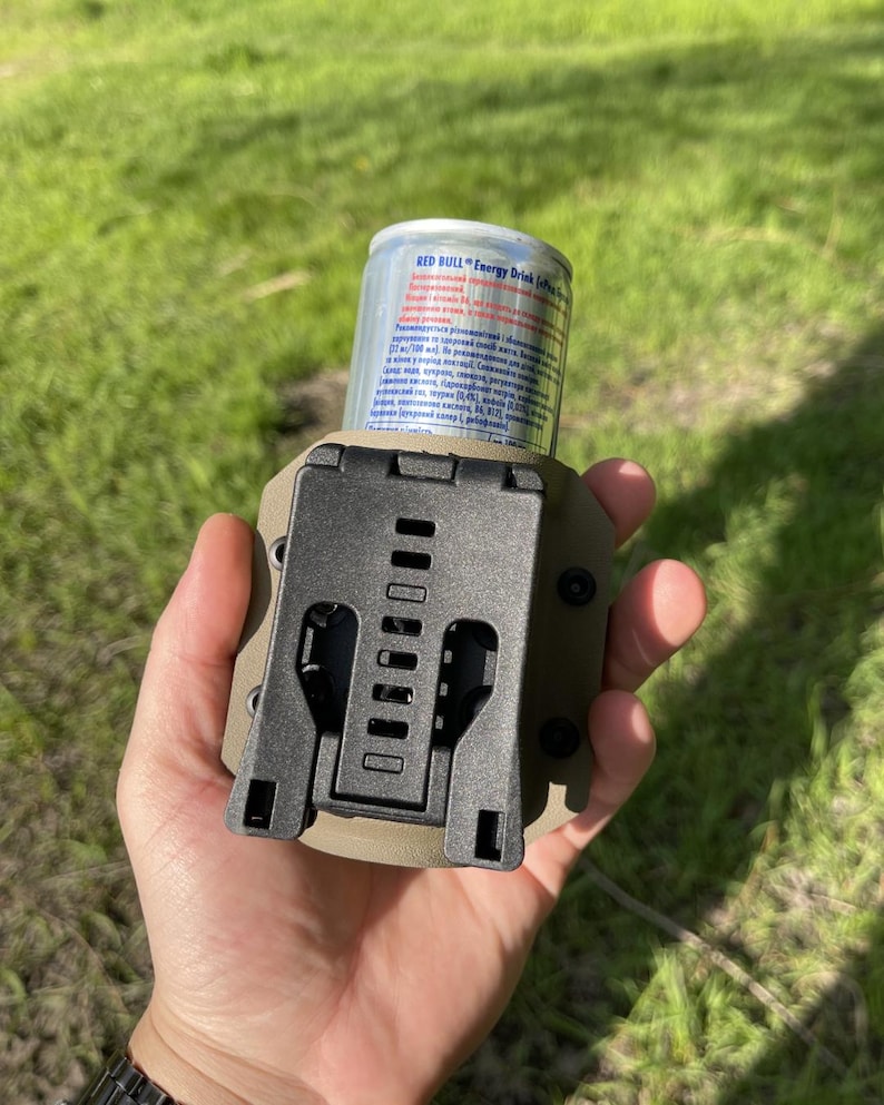 May include: A tactical beverage holder in olive green and black, designed to securely hold a Red Bull energy drink can. The holder features a sturdy, adjustable clip and is held in a hand against a blurred green background.