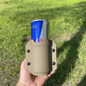 May include: A tan-colored drink holder with four black screws, holding a blue and silver can. The can has the Red Bull logo. The holder is made of a durable material and is designed to securely hold a beverage.