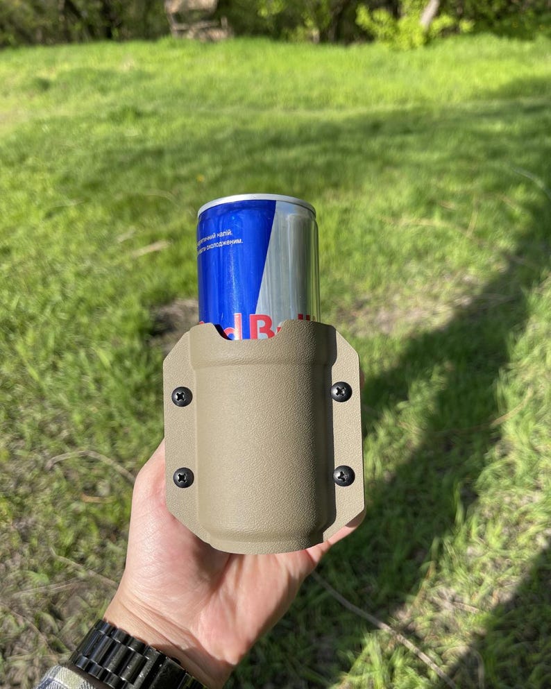 May include: A tan-colored drink holder with four black screws, holding a blue and silver can. The can has the Red Bull logo. The holder is made of a durable material and is designed to securely hold a beverage.