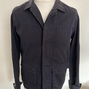 May include: A dark navy blue jacket with a collared neckline, long sleeves, and two front pockets. The jacket is displayed on a mannequin and has a button closure. The cuffs are rolled up.