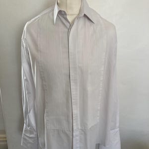 May include: A white dress shirt with vertical pleats down the front. The long-sleeved shirt is displayed on a mannequin. The shirt has a classic collar and buttoned cuffs. The fabric appears to be a crisp, lightweight material.