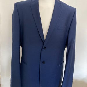 Könnte beinhalten: Ein marineblauer Blazer, der auf einer Schaufensterpuppe präsentiert wird. Der Blazer hat einen Zwei-Knopf-Verschluss, ein fallendes Revers und eine Brusttasche. Die Ärmel haben Knöpfe an den Manschetten. Der Blazer ist einfarbig und scheint aus einem glatten Stoff zu sein.