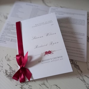 May include: A white wedding ceremony program with a burgundy ribbon tied in a bow. The program includes the names Shauna Wilson and Richard Ryan, the date Saturday, 26th July, 2025, and the location St. Brigid's Church.