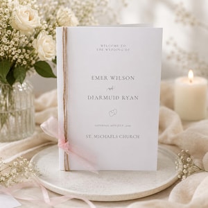 May include: A white wedding program with text that reads "WELCOME TO THE WEDDING OF EMER WILSON DIARMUID RYAN" and "SATURDAY, 26TH JULY 2025 ST. MICHAELS CHURCH". The program is tied with twine and a pink ribbon. A bouquet of white roses and a lit candle are in the background.