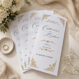 May include: White wedding programs with gold floral accents and elegant script. The programs read "Welcome to the Wedding of Catherine and Kierlliam, Saturday, 25th July, 2025, St. Michael's Church."
