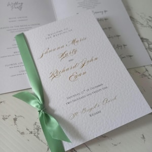 May include: Wedding ceremony program with a green ribbon. The program is white with gold lettering and includes the names Shauna Marie Harty and Richard John Egan, the date Saturday, 27th of December, Two Thousand and Twenty Five, and the location St Brigid's Church, Kildare.