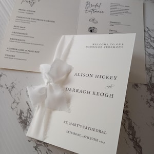May include: Wedding ceremony program with a white ribbon. The program includes the names of the bride and groom, the wedding party, and the ceremony details. The text is in black on an off-white background.
