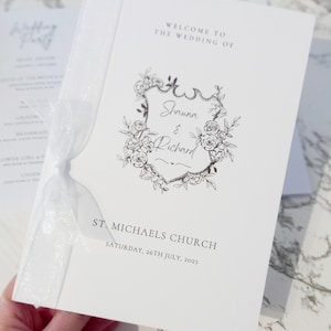 Puede incluir: Un programa de boda blanco con un diseño de escudo floral. El programa dice "Bienvenida a la boda de Shauna & Richard" y "St. Michael's Church, sábado 26 de julio de 2025". Una cinta blanca está atada alrededor del programa.