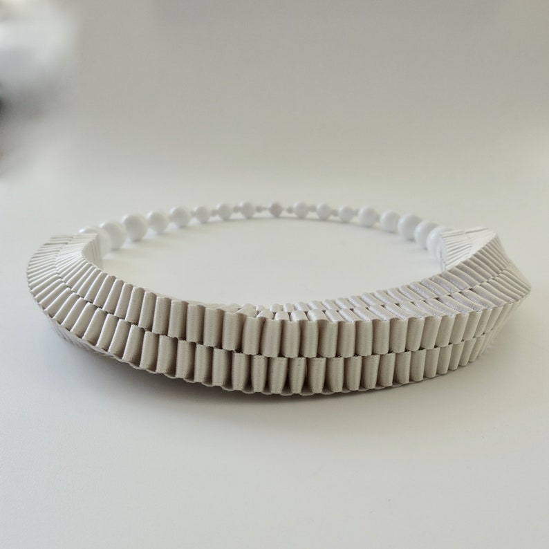 May include: A white, geometric necklace with a layered, textured design. The necklace features a series of vertical, ridged elements that create a unique, architectural look. Small, round beads form the back of the necklace, adding a delicate contrast.