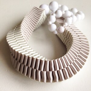 May include: A decorative necklace featuring a pleated, ribbon-like design in a soft beige color. The necklace is adorned with a strand of white, spherical beads. The overall aesthetic is elegant and minimalist.