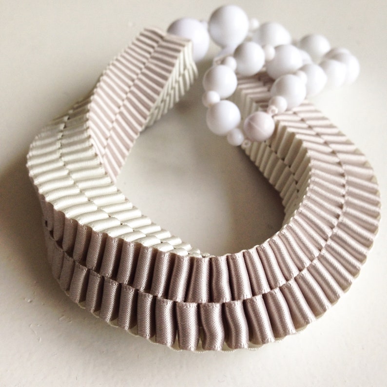 May include: A decorative necklace featuring a pleated, ribbon-like design in a soft beige color. The necklace is adorned with a strand of white, spherical beads. The overall aesthetic is elegant and minimalist.