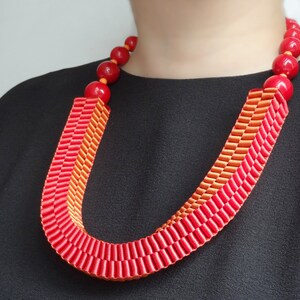 May include: A vibrant red and orange statement necklace. The necklace features large, round, red beads and a U-shaped design made of layered ribbons in shades of red and orange. The necklace is a bold accessory.