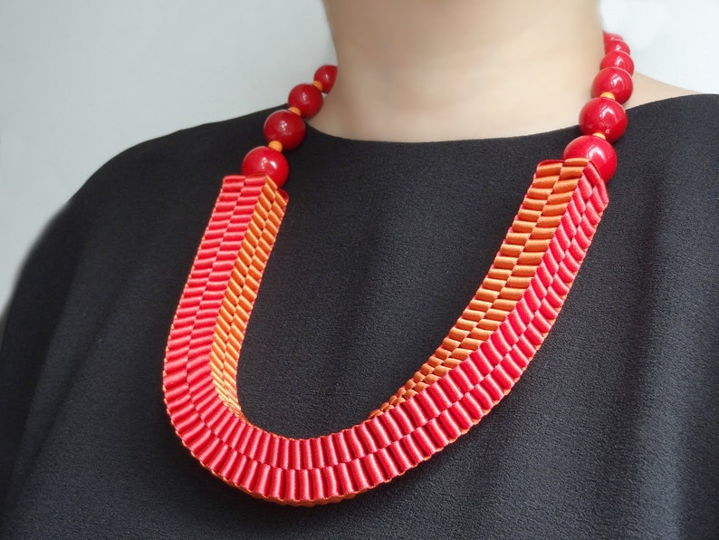 May include: A vibrant red and orange statement necklace. The necklace features large, round, red beads and a U-shaped design made of layered ribbons in shades of red and orange. The necklace is a bold accessory.