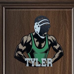 Puede incluir: Una silueta de lucha libre verde y negra con un protector de cabeza y el nombre "TYLER" impreso debajo. La silueta est&aacute; en una puerta de madera marr&oacute;n.