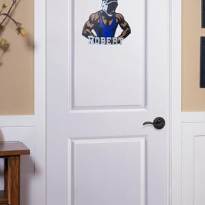 Puede incluir: Una puerta blanca con una calcoman&iacute;a de lucha libre que presenta a un luchador con un singlet azul y un protector de cabeza. La calcoman&iacute;a incluye el nombre "ROBERT" en letras plateadas.