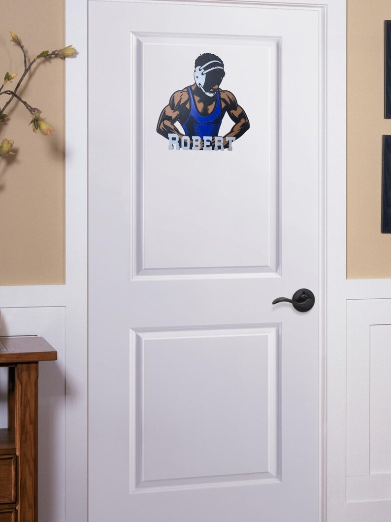 Puede incluir: Una puerta blanca con una calcoman&iacute;a de lucha libre que presenta a un luchador con un singlet azul y un protector de cabeza. La calcoman&iacute;a incluye el nombre "ROBERT" en letras plateadas.