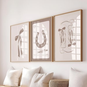May include: Three framed art prints with a western theme. The prints feature a bow, a horseshoe with the text "Lucky You", and cowboy boots. The prints are in neutral tones and are displayed above white storage cubes with pillows and a woven basket.