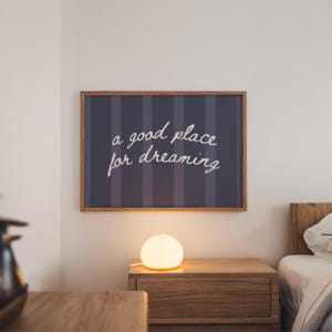 May include: A framed wall art piece with the text "a good place for dreaming" in white script. The artwork hangs above a wooden nightstand with a glowing spherical lamp. The background is a bedroom setting.