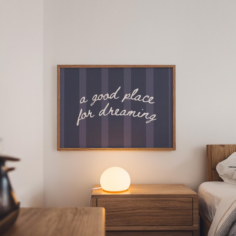 May include: A framed wall art piece with the text "a good place for dreaming" in white script. The artwork hangs above a wooden nightstand with a glowing spherical lamp. The background is a bedroom setting.