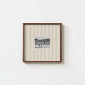 May include: A square wall frame with a dark brown wooden border and a light beige fabric mount featuring thin vertical stripes. The centre contains a black and white photograph of three figures standing on a beach near a large body of water.