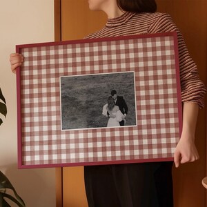 May include: A framed photograph with a black and white image of a couple. The frame is a deep red with a red and white gingham pattern. The frame is held by a person.