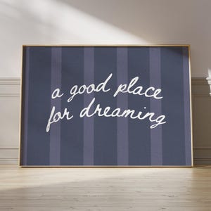 May include: A framed print with the words "a good place for dreaming" in white script on a navy blue striped background. The frame is gold, and the print is leaning against a wall.
