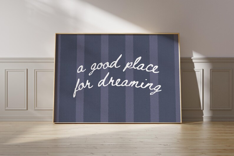 May include: A framed print with the words "a good place for dreaming" in white script on a navy blue striped background. The frame is gold, and the print is leaning against a wall.