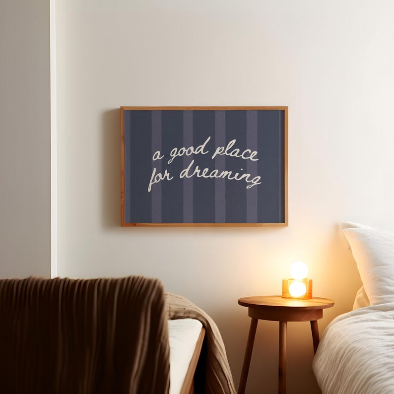 May include: A framed wall art piece with the text "a good place for dreaming" in white script. The artwork has a navy blue background with vertical stripes and a wooden frame. The scene includes a bed, a side table, and a lamp.