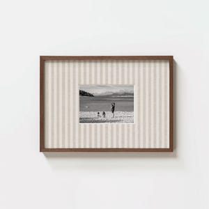 May include: A rectangular picture frame with a dark brown wooden border and a beige and white vertical striped mat. The frame contains a black and white photograph of a beach scene with three figures standing near the water and mountains.