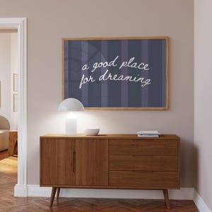 May include: A wooden mid-century modern style sideboard with a framed print above it. The print has the words "a good place for dreaming" in white script. A white lamp and bowl sit on top of the sideboard.