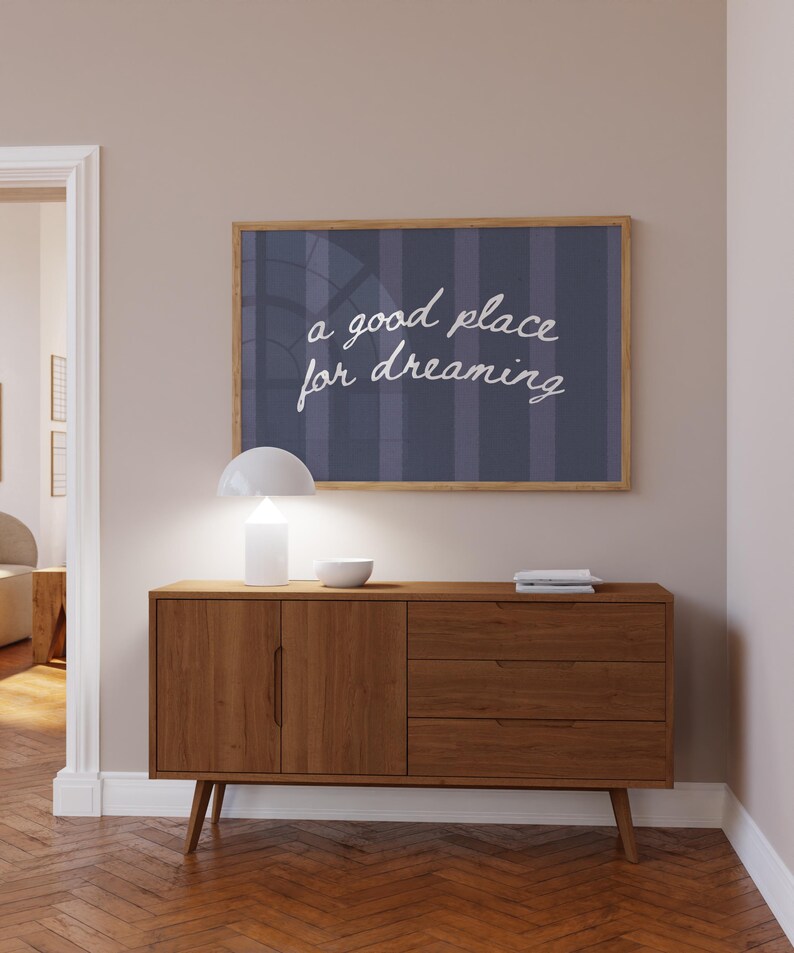 May include: A wooden mid-century modern style sideboard with a framed print above it. The print has the words "a good place for dreaming" in white script. A white lamp and bowl sit on top of the sideboard.