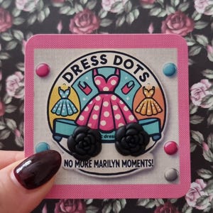 May include: A pair of black rose-shaped earrings with a pink and white card that says "Dress Dots" and "No More Marilyn Moments!" The card features a cartoon illustration of three dresses.