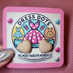 May include: A pair of heart-shaped earrings in a light brown color. The earrings are displayed on a pink and white card with the text "Dress Dots" and "No More Marilyn Moments!"