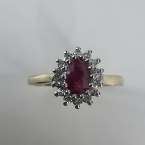 May include: A gold ring with a large, oval-cut, deep red gemstone encircled by a halo of smaller, clear gemstones. The ring's band is plain, polished gold.