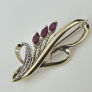 Puede incluir: Un broche de oro con un diseño delicado y curvo. El broche está adornado con tres piedras de rubí rojas y una fila de piedras blancas más pequeñas.