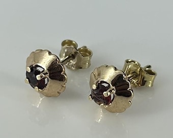 May include: A pair of gold stud earrings with a small, round, dark red gemstone in the centre of each earring.