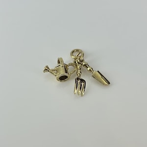 May include: A set of three gold-toned metal charms featuring a miniature watering can, a small garden fork, and a trowel. Each charm is equipped with a small circular loop at the top for attachment, showcasing detailed gardening tool designs.