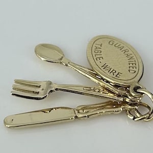 Könnte beinhalten: Ein goldfarbener Anhänger mit Miniatur-Besteck: eine Gabel, ein Löffel und ein Messer. Ein angehängtes Etikett trägt die Aufschrift "GUARANTEED TABLE WARE". Der Anhänger ist wahrscheinlich für eine Halskette oder ein Armband.