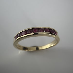 May include: A gold ring featuring a line of round, deep red gemstones. The ring has a smooth, polished surface and a gently curved shape. The gemstones are evenly spaced and set into the band.