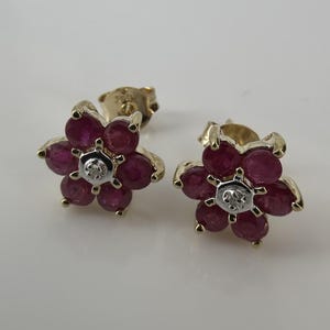 Peut inclure: Une paire de boucles d'oreilles à tige en forme de fleur en or jaune. Chaque boucle comporte cinq rubis ronds rose foncé disposés en pétales autour d'une petite pierre centrale ronde et transparente sertie dans du métal blanc.