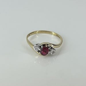 May include: A gold ring featuring a central oval-cut red gemstone flanked by two small round-cut clear stones. The setting for the stones is silver-toned, contrasting with the smooth, polished yellow gold band that tapers toward the back.