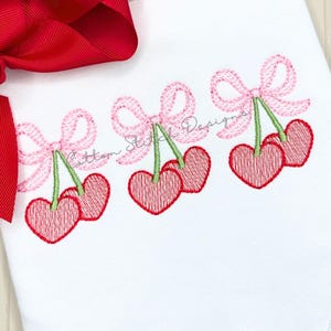 May include: White fabric with three embroidered cherry designs. Each cherry design features two red heart-shaped cherries, green stems, and a pink bow. A large red bow is visible in the upper left corner.